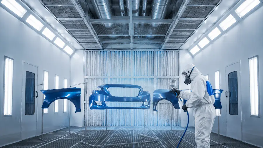 Cabine de pintura automotiva com sistema de filtragem de ar no teto, peças automotivas suspensas e profissional realizando pintura com equipamento de proteção.